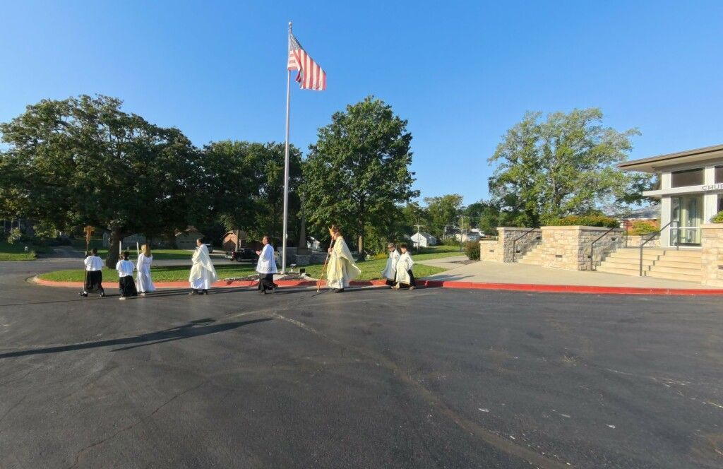 A group of people in white and light-colored robes walk past an American flag on a sunny day. 