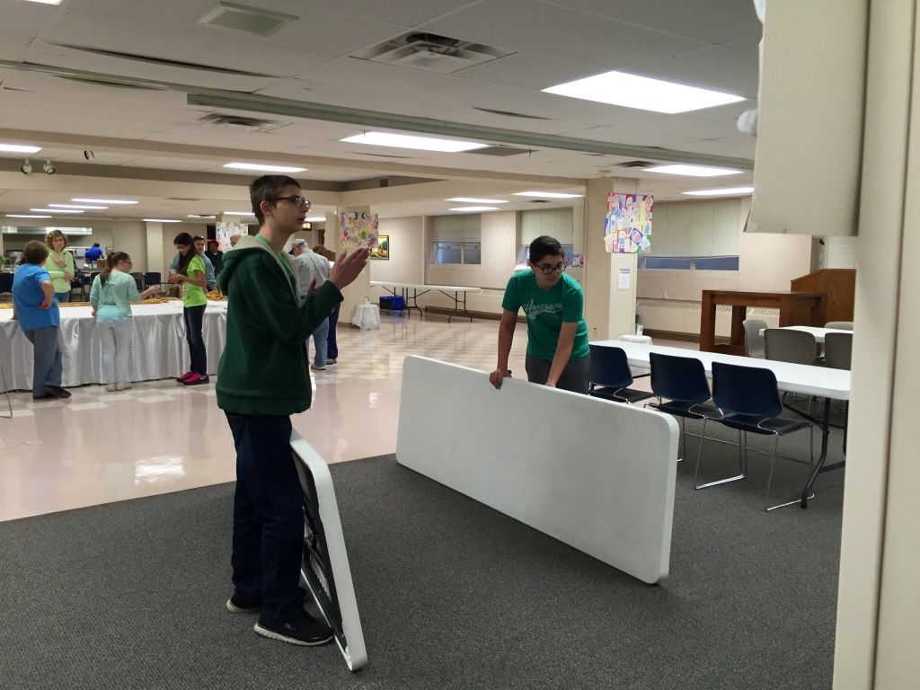 Two people set up tables in a spacious room with tiled floors and fluorescent lighting. 