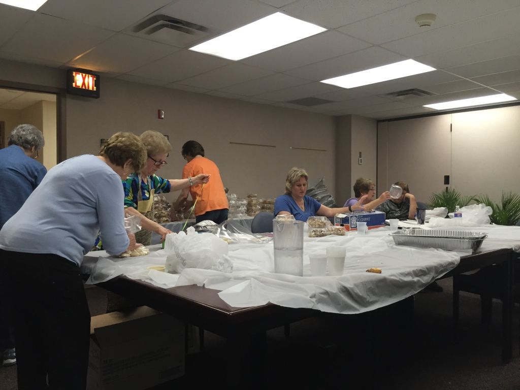 A group of people in a room are preparing food, surrounded by trays and bags.