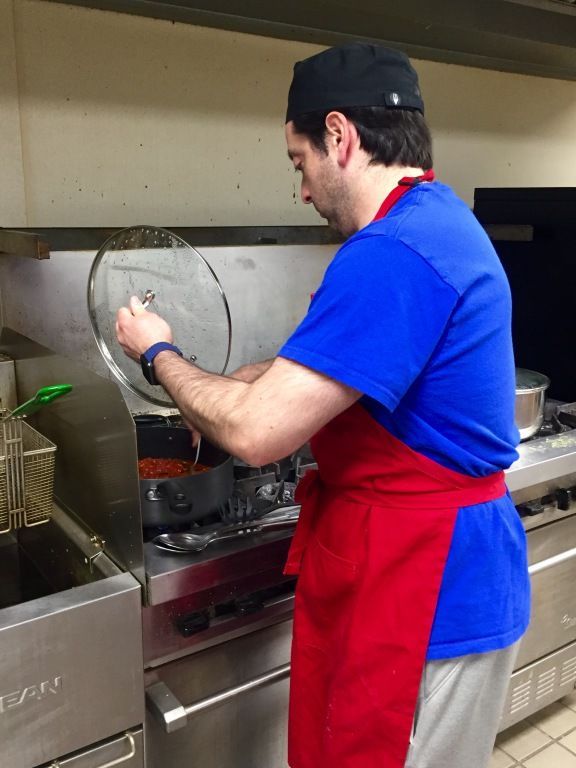 A man in a blue shirt and red apron lifts a pot lid while cooking in a commercial kitchen.