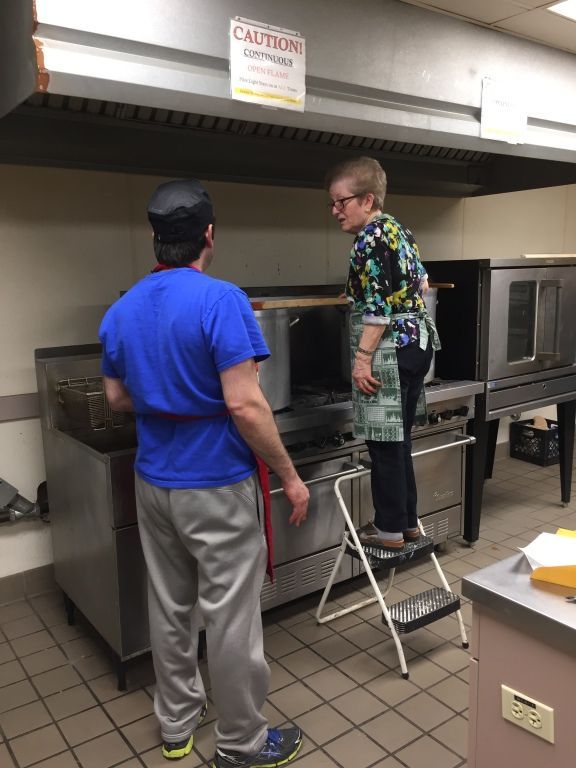 Two people in a kitchen, one on a step stool tending to a stove.