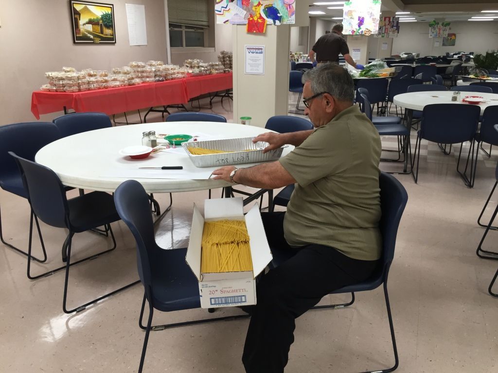 A man sits at a round table in a community hall, organizing pasta into a large aluminum tray. 