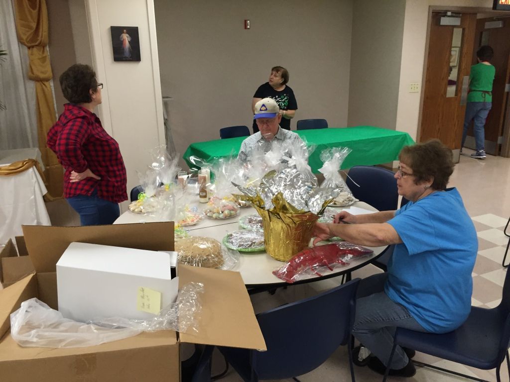 Three people are gathered around a table preparing gift baskets with plastic wrap and decorations.