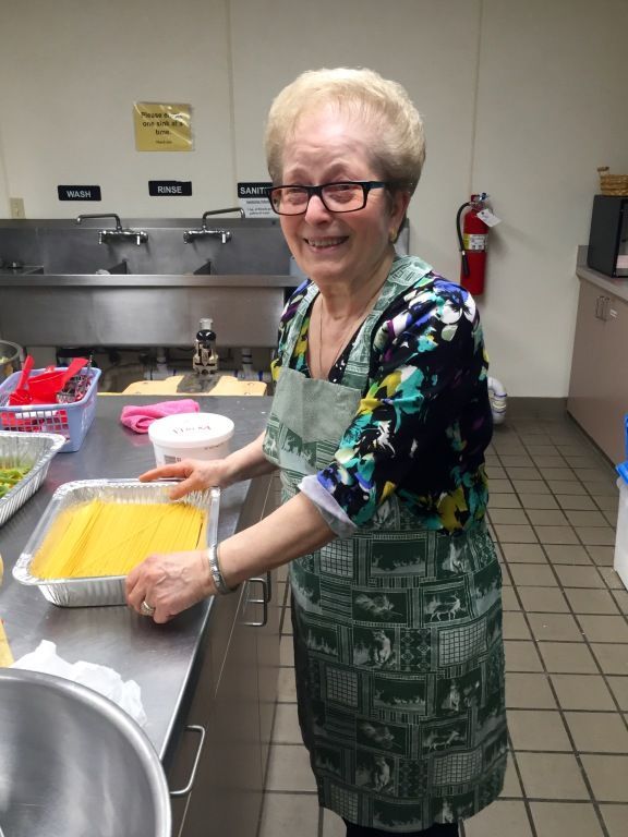 Elderly woman with glasses, smiling in a kitchen, preparing pasta in a tray.