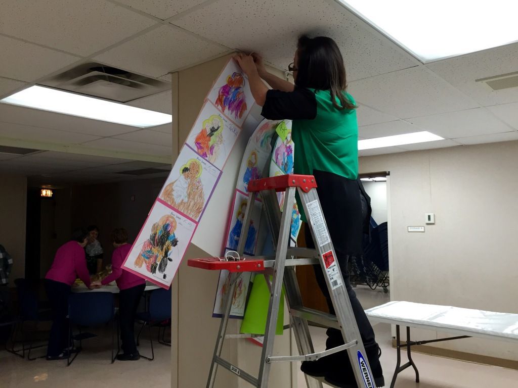 A woman in a green top stands on a ladder, attaching colorful artwork to a wall in a well-lit room.