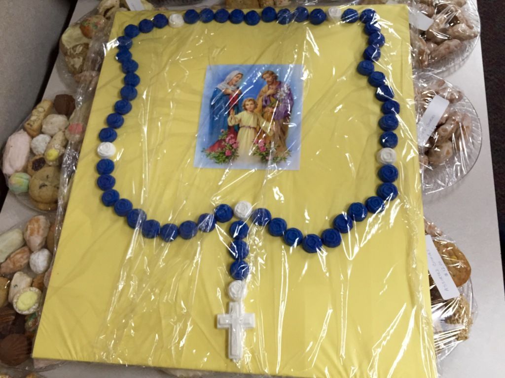 A decorative cake with a yellow surface, featuring a blue and white rosary design and a small image of the Holy Family at the center.