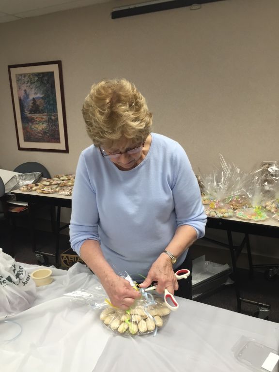 A person in a light blue sweater wraps a tray of cookies with clear cellophane on a white table.