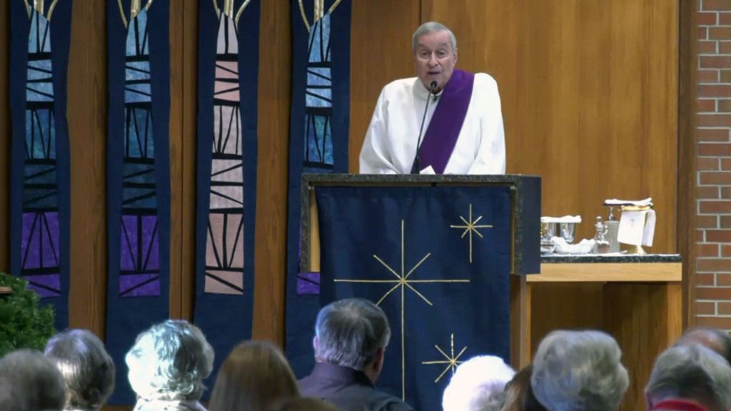 A clergy member in white and purple robes speaks at a church podium adorned with a star symbol.