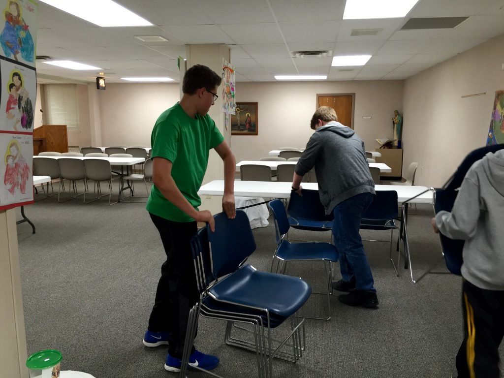 Three people arranging blue chairs in a room with white walls and tables. 