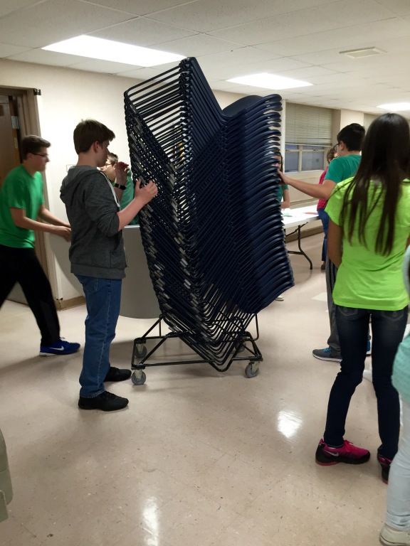 A group of people, some in green shirts, stack blue metal chairs on a wheeled rack in a bright room.