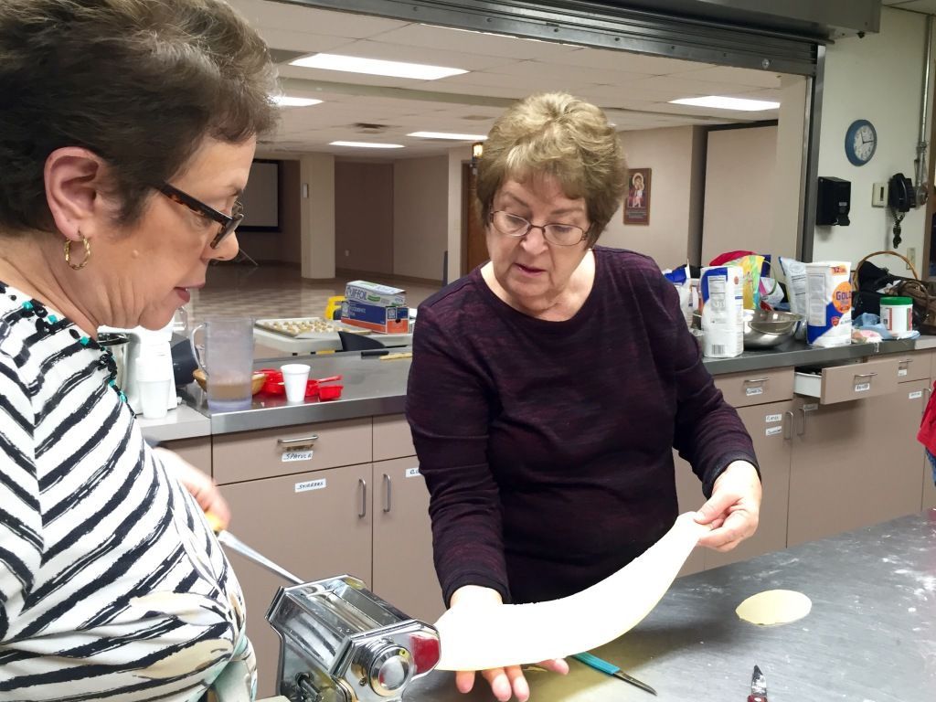 Two women work together in a kitchen, rolling pasta through a pasta maker.