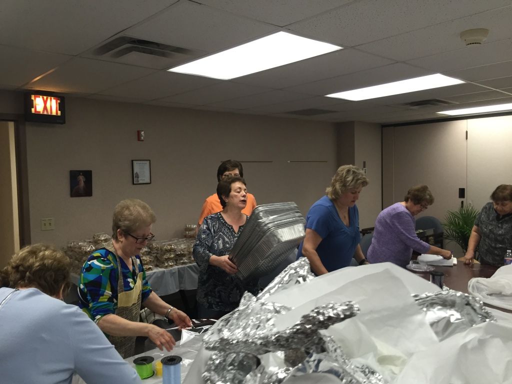 A group of women in a kitchen prepares food trays, surrounded by tables and foil. 