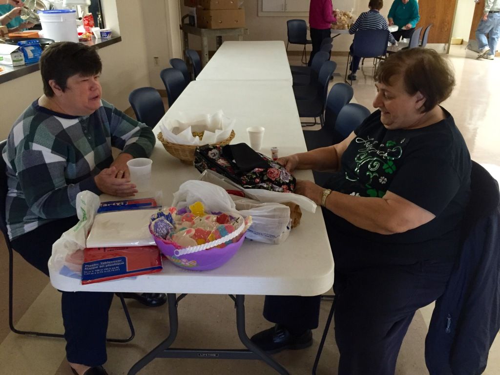 Two women sit at a long, white table in a communal space, chatting and smiling.
