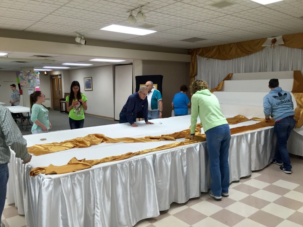 A group of people collaborate to set up a large banquet table covered with white and gold drapes in a hall.