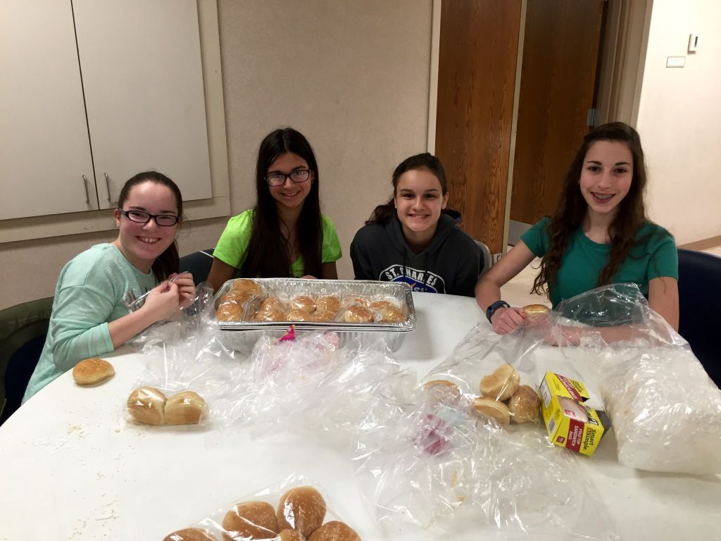 Four smiling girls sit around a table filled with bread rolls wrapped in plastic. 