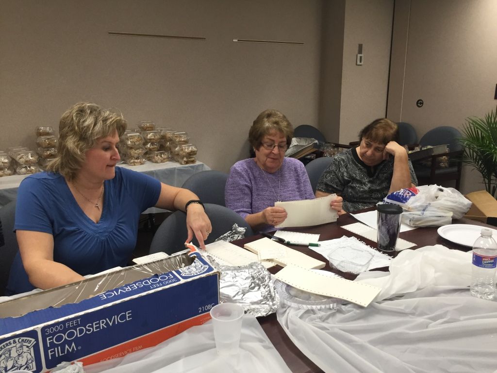 Three women sit around a table covered with papers, foil, and plastic wrap in a work setting.
