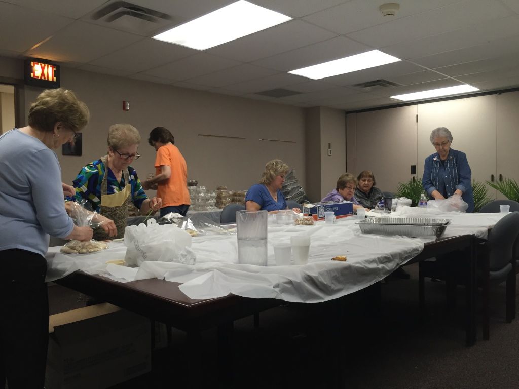 A group of women in a community center is busy preparing food at a table covered in a white cloth. 
