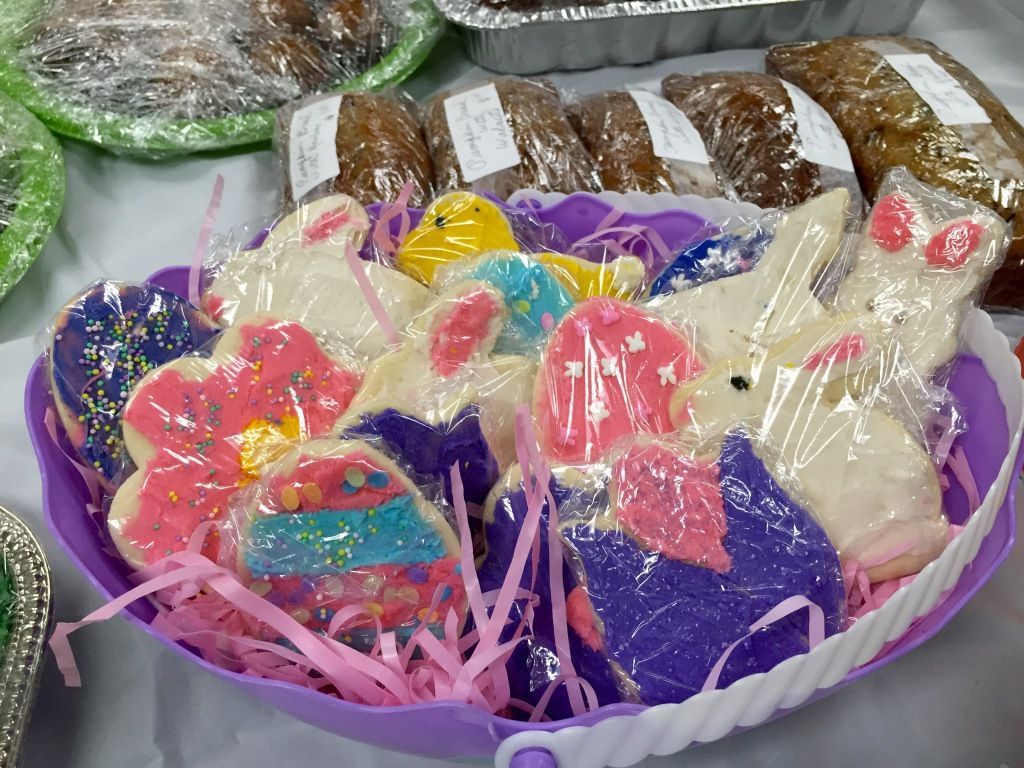 A purple basket holds colorful, icing-covered Easter cookies shaped like flowers and bunnies, wrapped in plastic.
