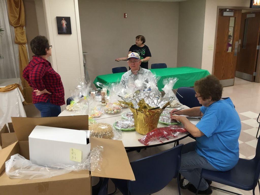 Three people at a round table prepare gift baskets wrapped in shiny paper.