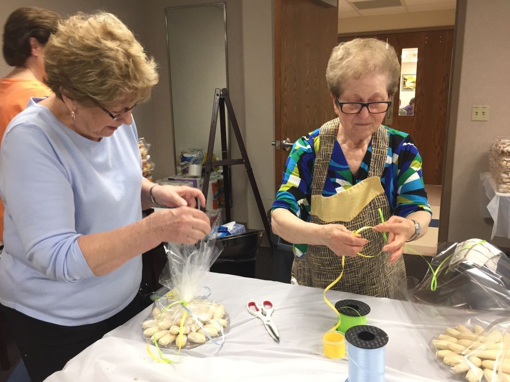 Two older women are packaging cookies in cellophane with colorful ribbons at a table.