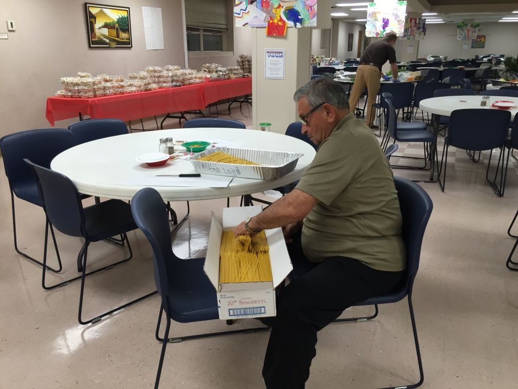 A man sits at a round table in a community hall, organizing pasta into a large aluminum tray. 