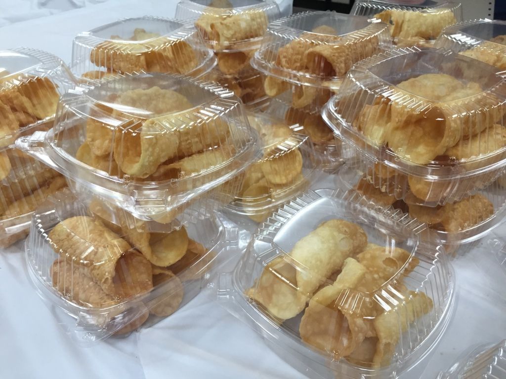 Stacks of clear plastic containers filled with golden, crispy cannoli shells are arranged on a white table.