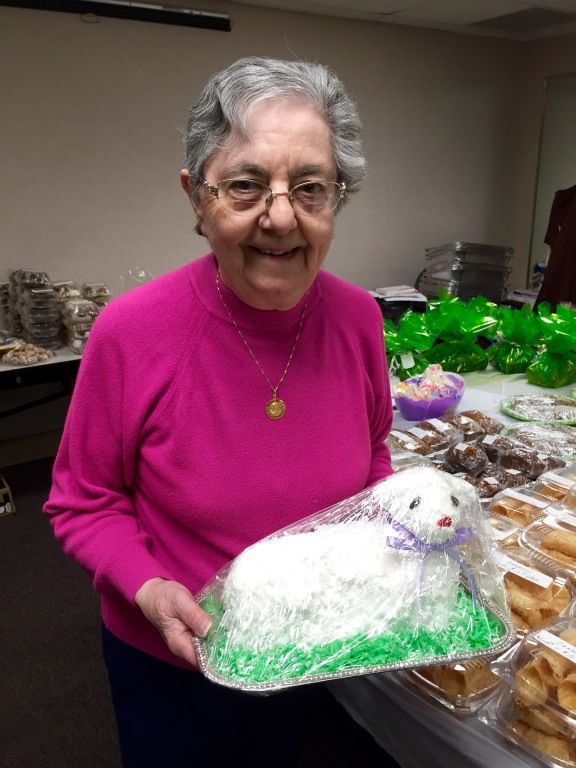 Elderly woman in a pink sweater smiles, holding a lamb-shaped cake wrapped in plastic. 