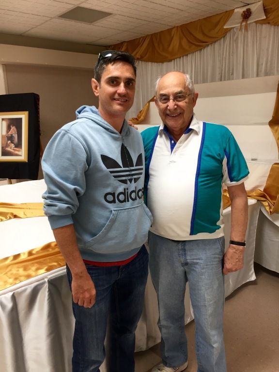 A smiling older man in a colorful polo shirt stands beside a younger man in a blue Adidas hoodie. 