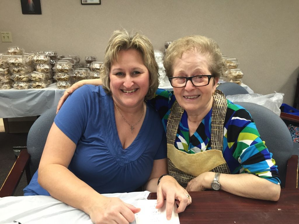 Two women smiling warmly, sitting close together in a room with stacks of packaged cookies.