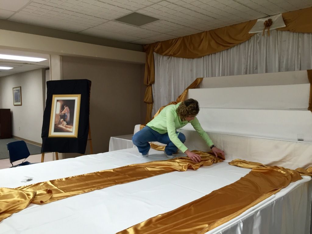 A person wearing a green shirt arranges gold fabric on a large, white-covered platform in a decorated room. 