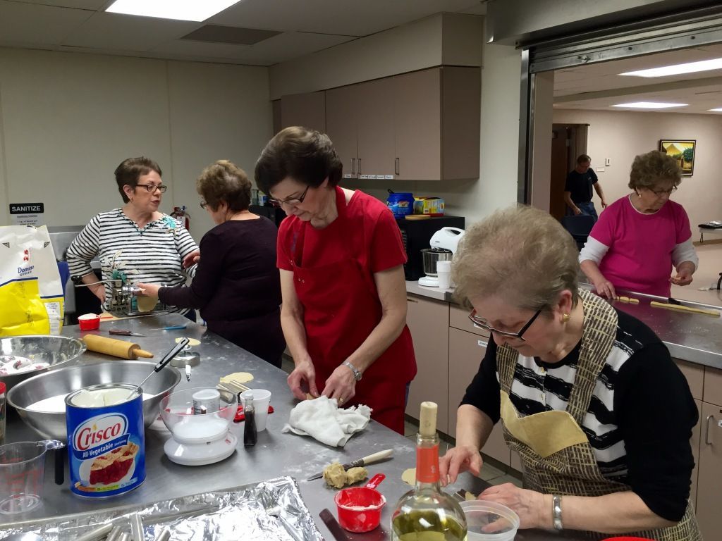 A group of five women are preparing food in a kitchen, wearing aprons.