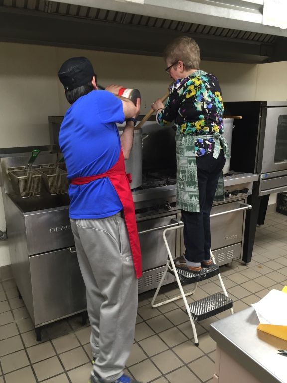 Two people in a commercial kitchen, one pouring from a large can while the other stirs a big pot on a stove. 