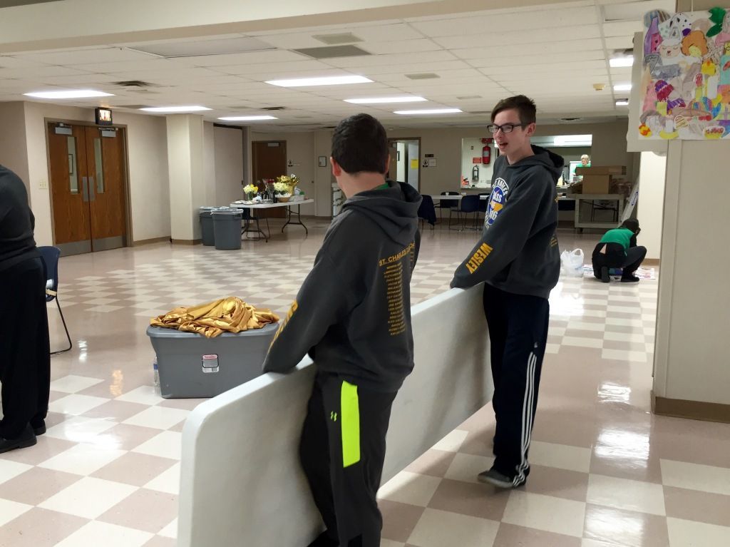 Two young people are carrying a large folded table in a hallway with a tiled floor.