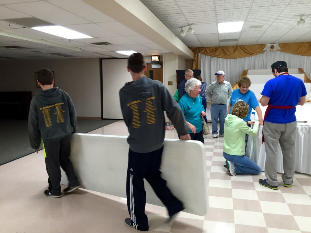 Two young men in matching hoodies carry a folding table in a community hall. 