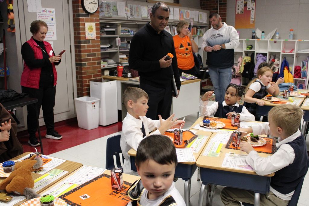 Children in school uniforms sit at desks with Halloween-themed snacks and drinks.