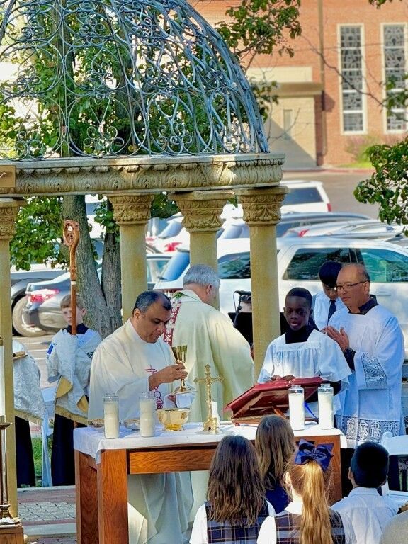 A priest in white robes conducts a religious ceremony outdoors, under a decorative gazebo.