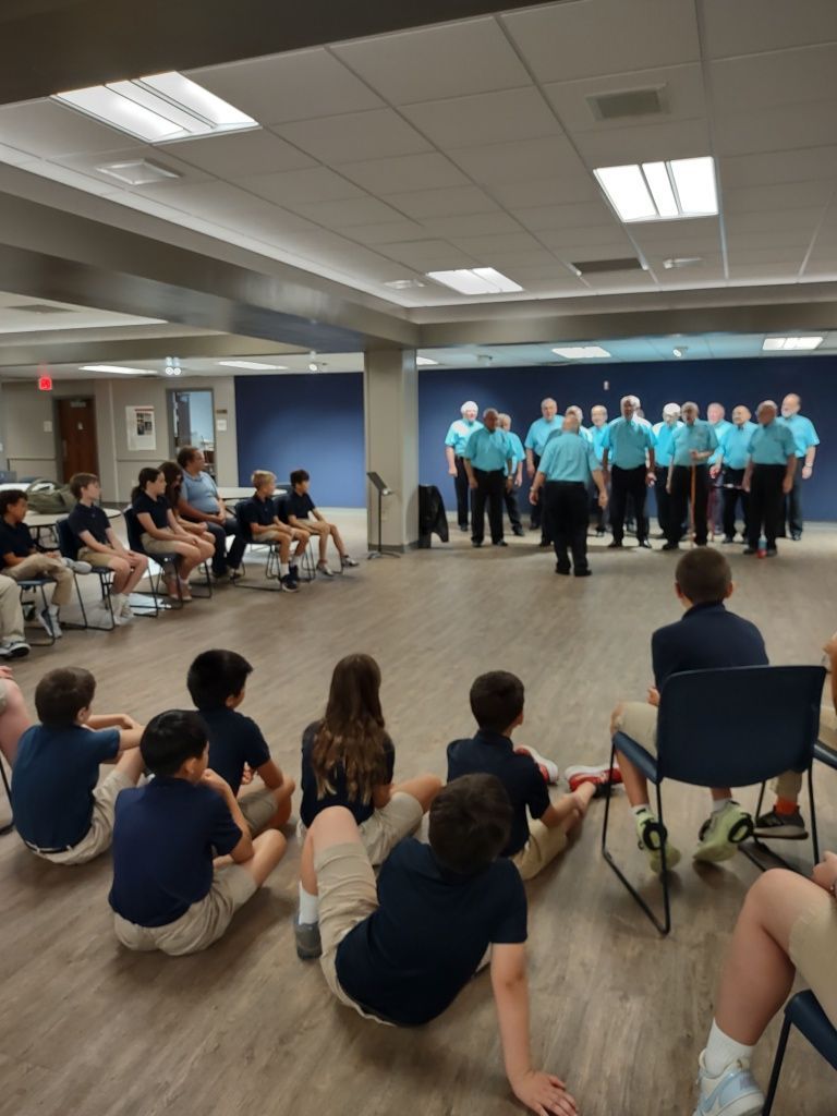 A group of singers in teal shirts performs passionately for seated children in a spacious room.