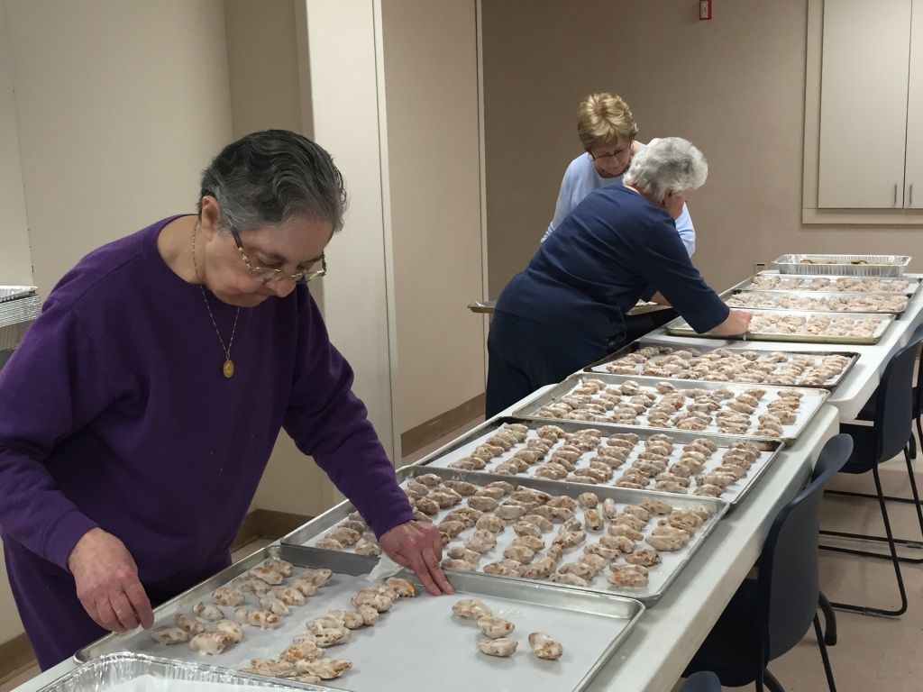 Three women are arranging trays of dough pieces on a long table in a room.
