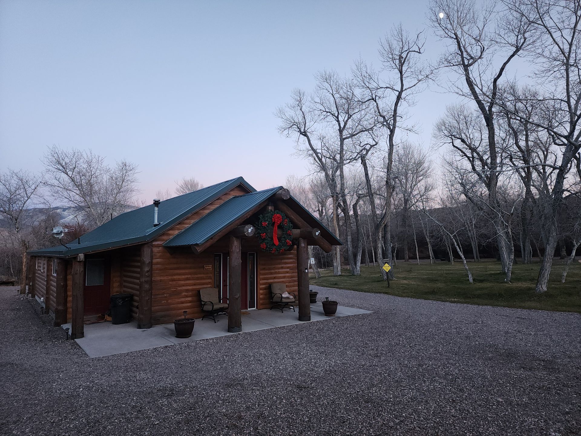 A rustic log cabin with a dark metal roof sits on a gravel lot surrounded by bare trees at twilight.