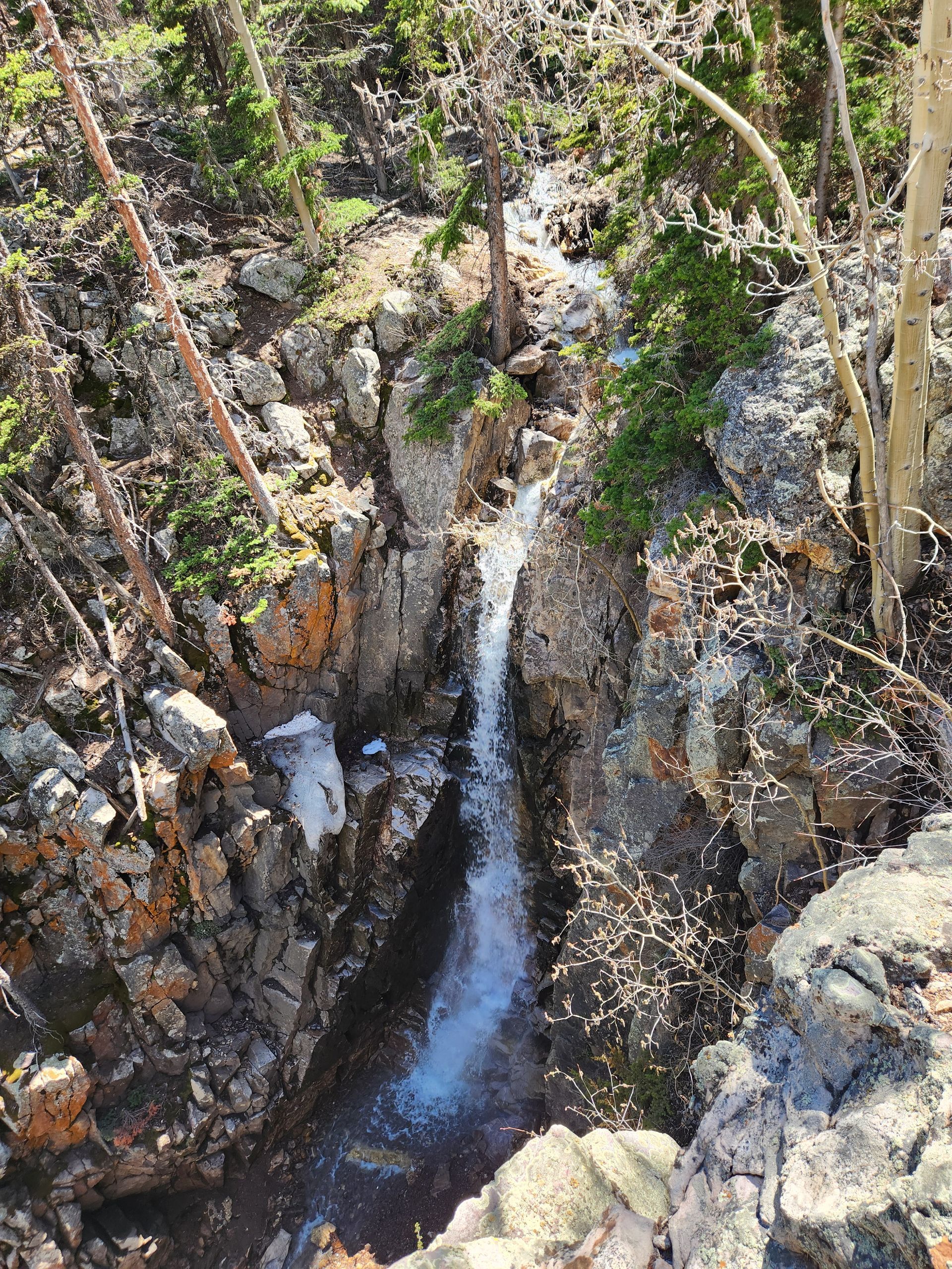 A small, narrow waterfall flows through a rocky crevice surrounded by trees and patches of snow in a forest.