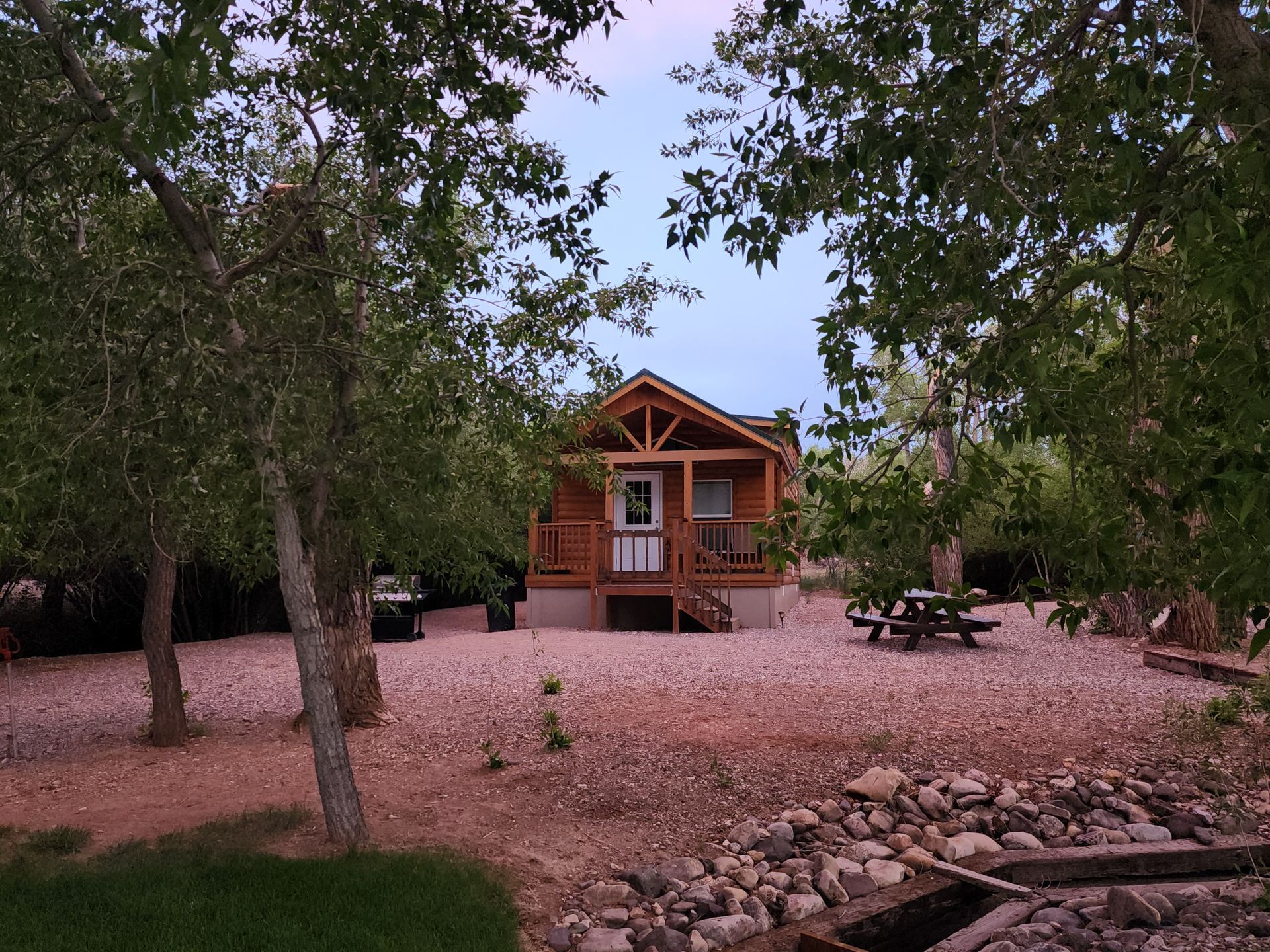 A small wooden cabin with a porch sits in a clearing with a picnic table, surrounded by trees and gravel ground.