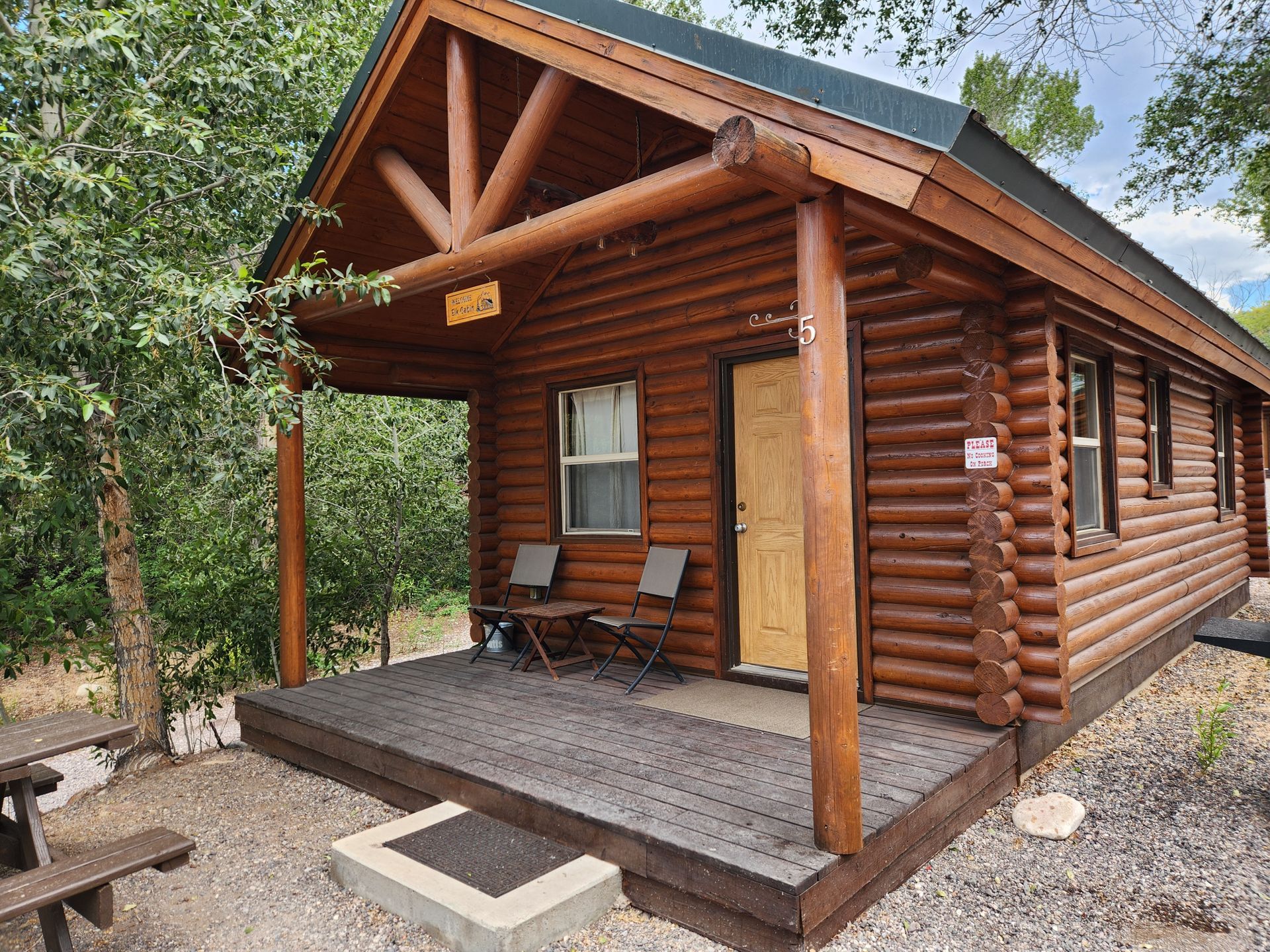 A rustic log cabin with a wooden porch, two chairs, and a metal roof, set in a wooded outdoor area.