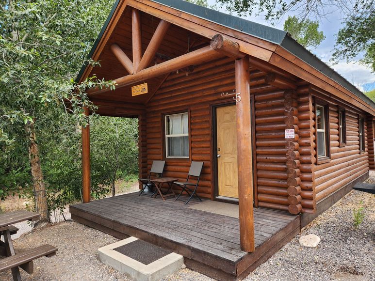 A rustic log cabin with a wooden porch, two chairs, and a metal roof, set in a wooded outdoor area.