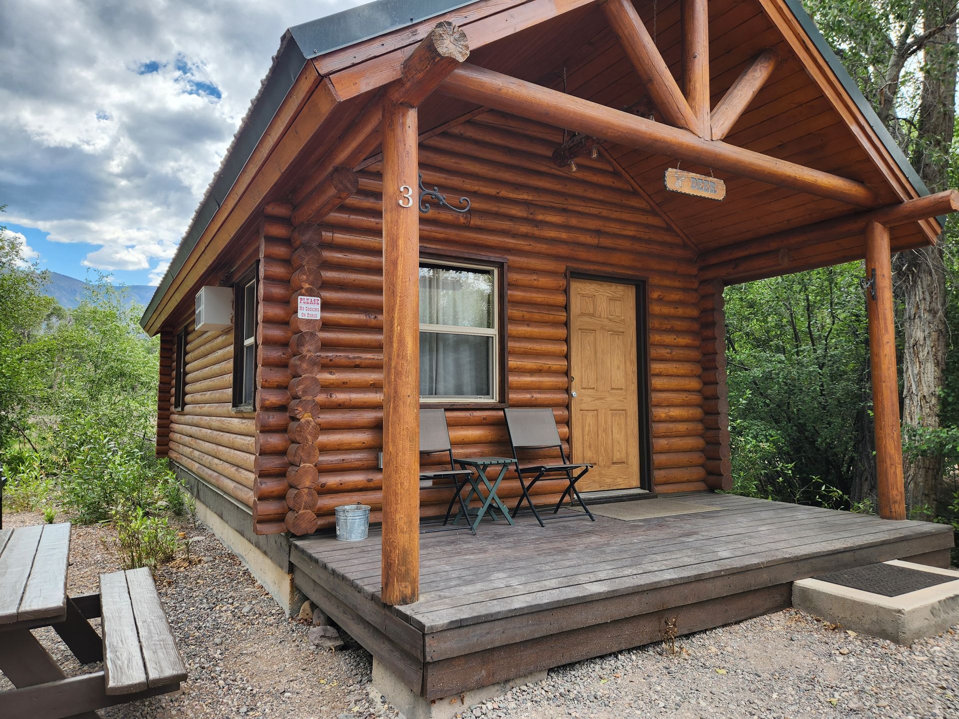 A small wooden log cabin with a covered front porch, two folding chairs, and a picnic table in a wooded, sunny setting.