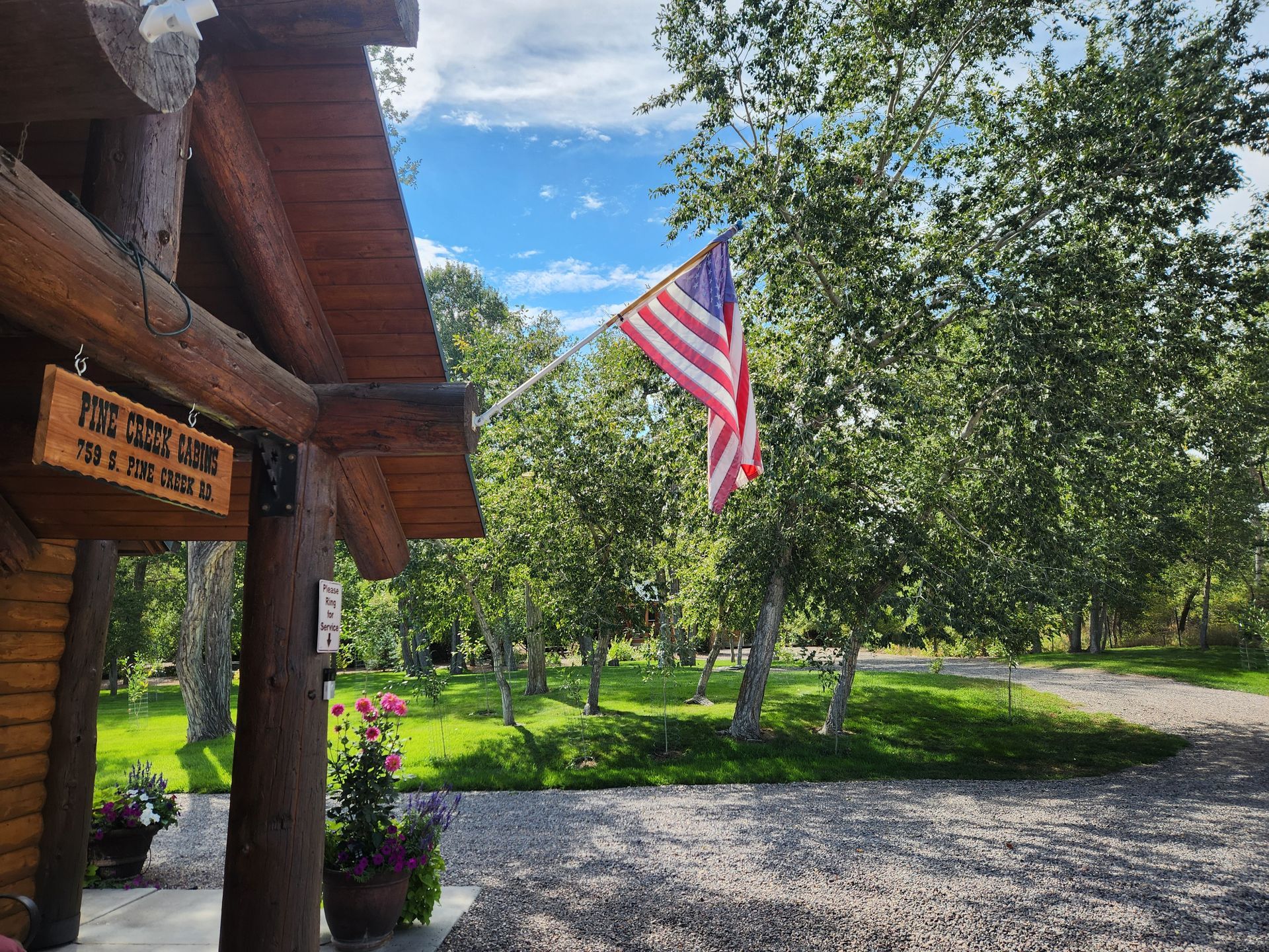 An American flag waves in front of a log cabin with a wooden sign, surrounded by green trees and a gravel driveway.