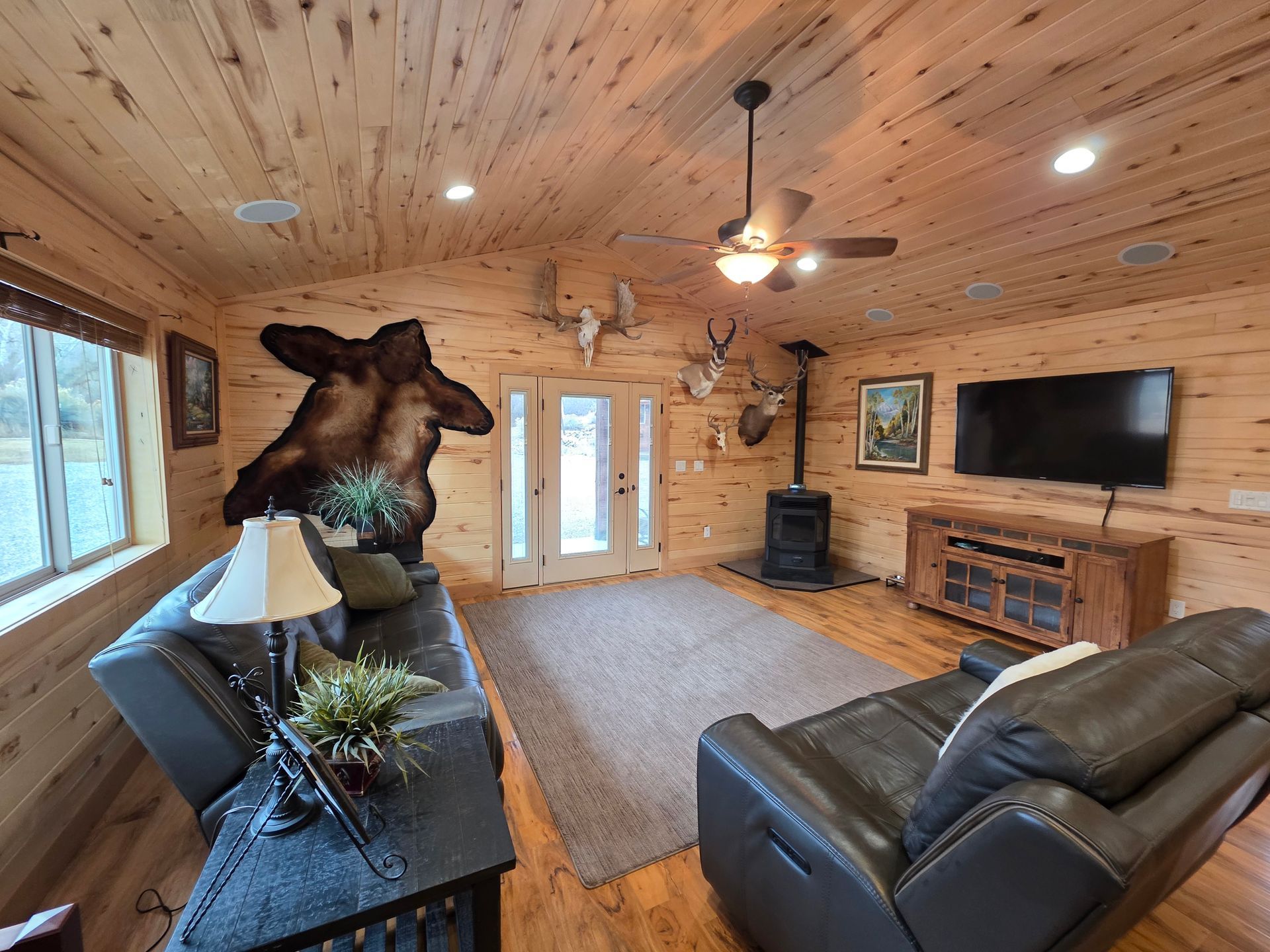A cozy living room with knotty pine walls, dark leather furniture, a TV, wood stove, and decorative wildlife taxidermy.