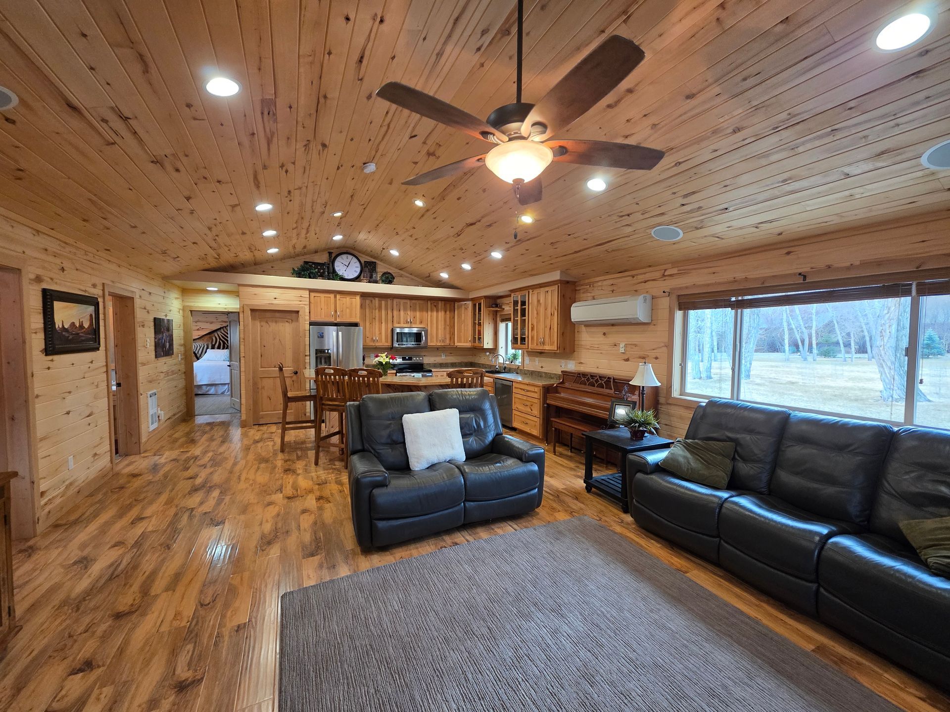 A living room and kitchen area featuring wood paneling, dark leather sofas, a ceiling fan, and a gray area rug.