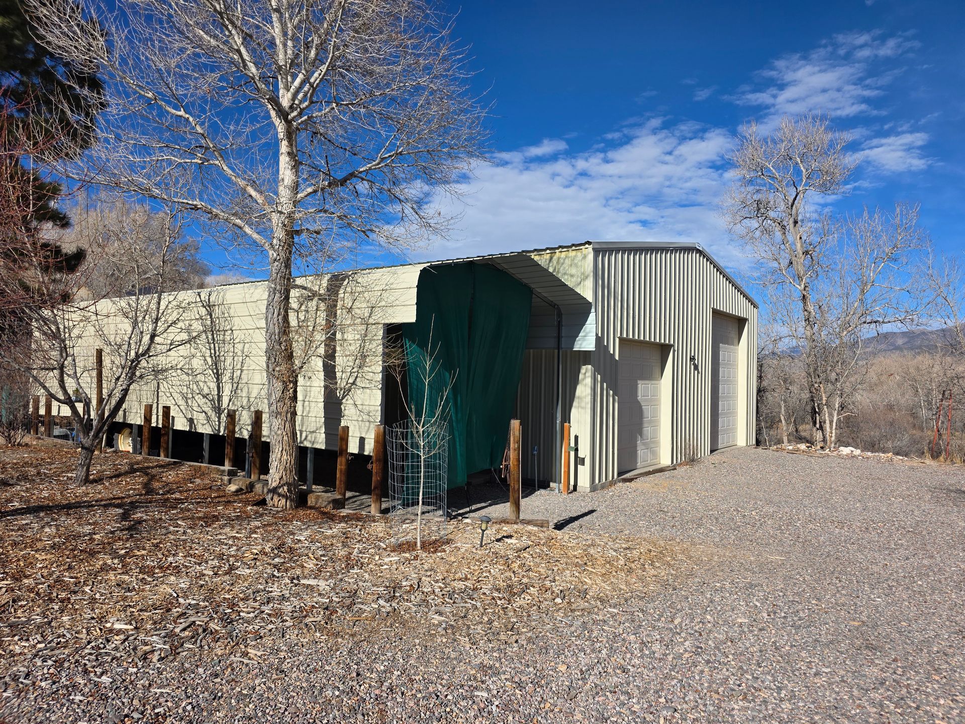 A beige metal building with two garage doors and a green side tarp sits on a gravel lot under a bright blue sky.