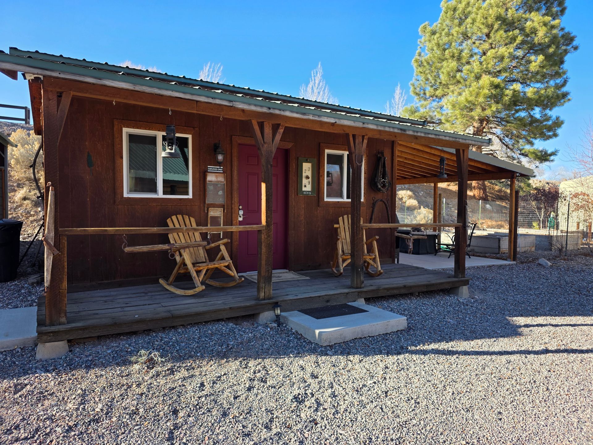 A rustic brown wooden cabin with a covered front porch featuring two rocking chairs, set on a gravel lot under blue sky.