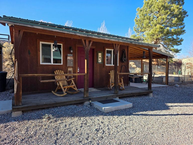 A rustic brown wooden cabin with a covered front porch featuring two rocking chairs, set on a gravel lot under blue sky.